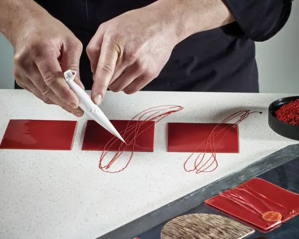 Mains d'un pâtissier créant un décor royal chocolat sur des carrés rouges brillants.