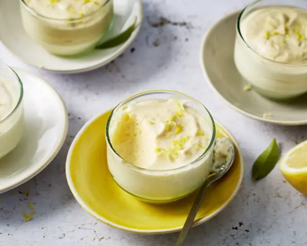 Plusieurs verrines de mousse au citron, garnies de zestes, sur des assiettes claires et jaunes. Un citron coupé est visible à droite.