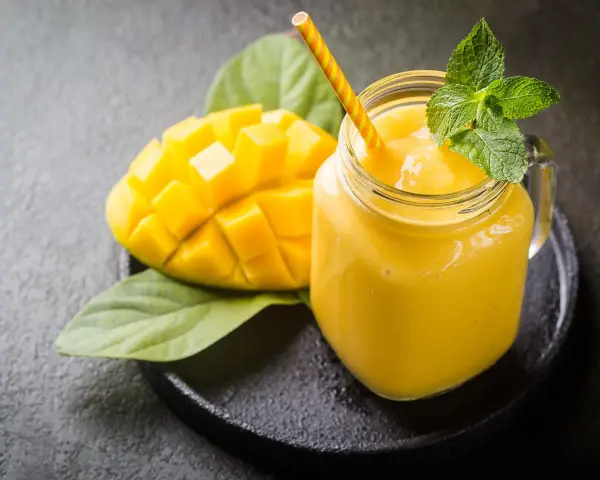 Un smoothie mangue onctueux dans un pot en verre, garni de menthe fraîche, à côté d'une moitié de mangue coupée en dés.