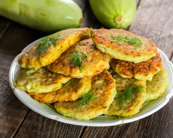Pile de beignets de courgette dorés et moelleux, garnis de brins d'aneth frais, sur une assiette blanche. Deux courgettes vertes floues en arrière-plan.