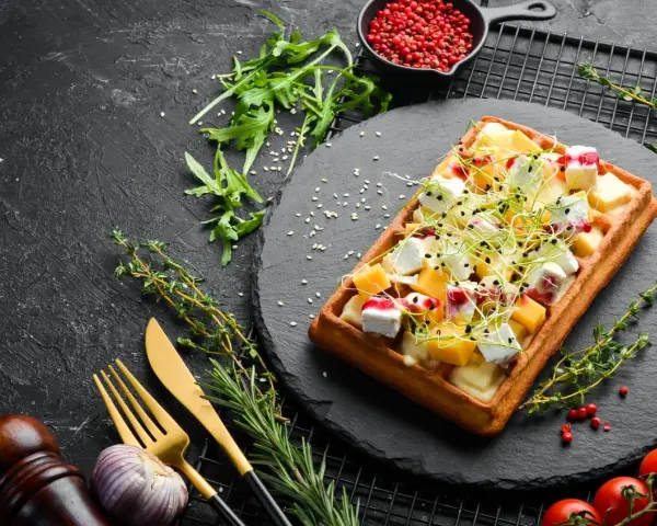 Gaufre salée garnie de fromage, de dés de mangue et de pousses, sur une ardoise noire.