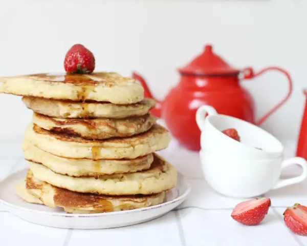 Pile de pancake sans gluten, nappés de sirop et garnis d'une fraise. Une théière rouge et des tasses complètent la scène.