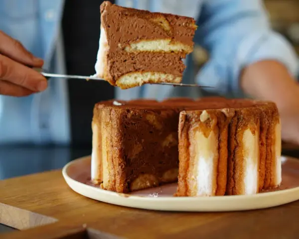 Une part de charlotte tiramisu, aérienne et chocolatée, est prélevée d'un gâteau entier.