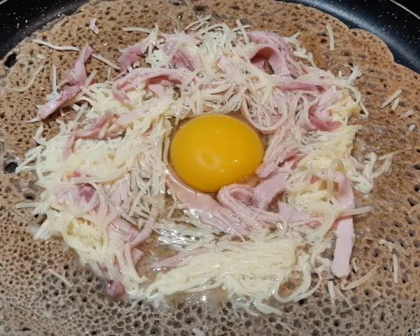 Une galette sarrasin garnie d'un œuf, de jambon et de fromage râpé, prête à être cuite dans une poêle.