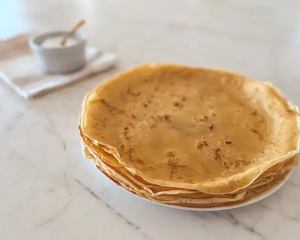 Pile de crêpes dorées, parfaites pour une recette crêpe moelleuse. Un petit bol de sucre est à côté.
