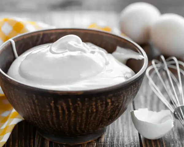Un bol rempli de crème fouettée, blanc en neige, prêt à être utilisé pour une pâtisserie. Des œufs et un fouet sont visibles à proximité.