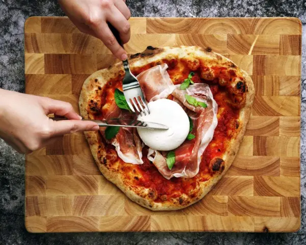 Une pizza burrata avec du prosciutto et du basilic, prête à être dégustée. Des mains coupent la burrata crémeuse.