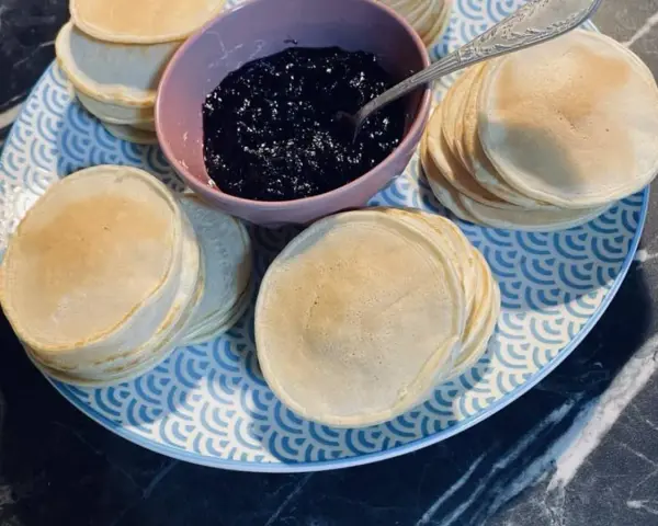 Une assiette bleue et blanche présente des piles de poffertjes dorés, accompagnés d'un bol de confiture de myrtilles. Parfait pour une recette de poffertjes !