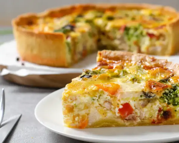 Une part de quiche savoureuse avec une pâte à tarte maison dorée, garnie de légumes et de poulet. Le reste de la tarte est flou en arrière-plan.