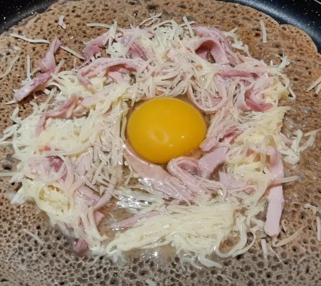 Une galette sarrasin garnie d'un œuf, de jambon et de fromage râpé, prête à être cuite dans une poêle.