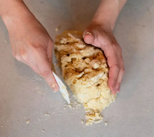 Mains travaillant une pâte sablée, la façonnant avec une spatule sur un plan de travail.