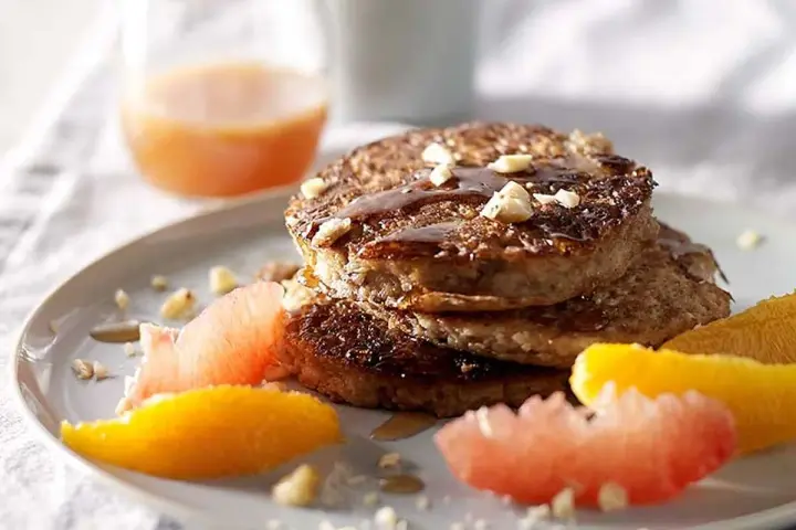 Pile de pancakes muesli, nappés de sirop et garnis de noix, accompagnés de quartiers d'agrumes.