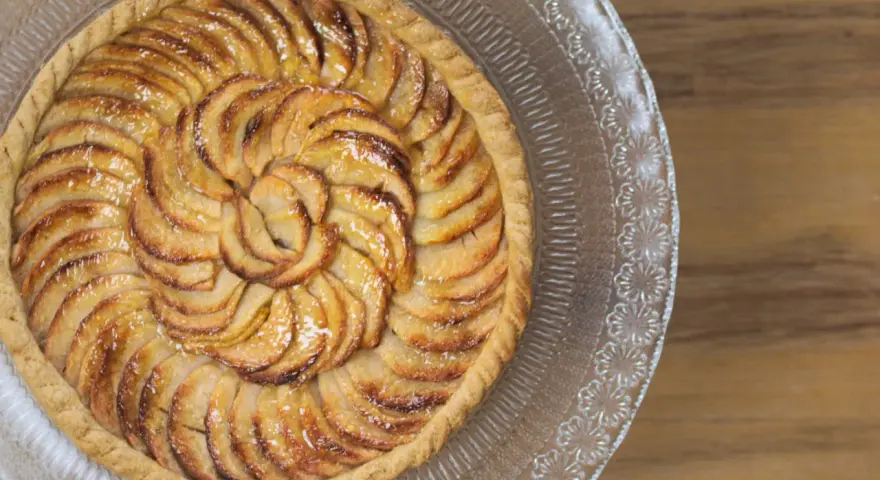 Un magnifique appareil tarte aux pommes, aux pommes caramélisées disposées en spirale sur une pâte dorée, sur un plat en verre décoré.