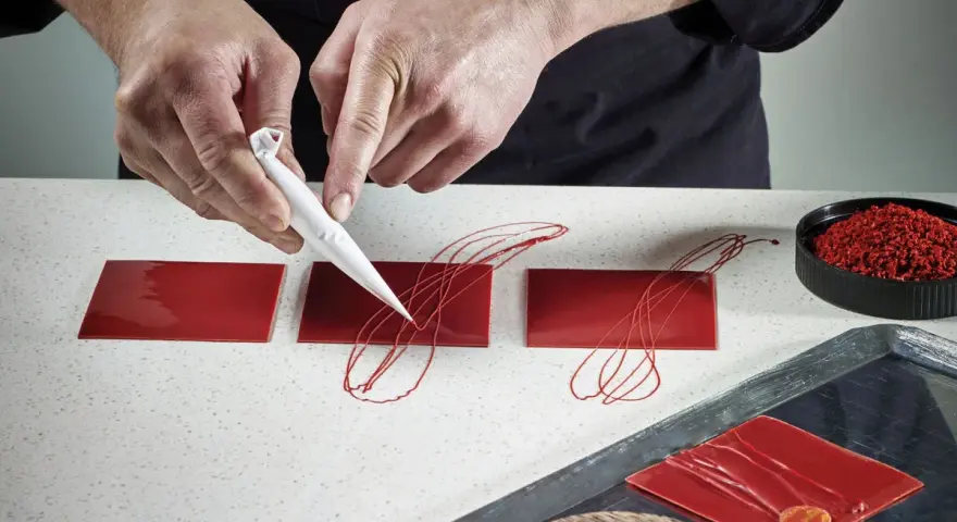 Mains d'un pâtissier créant un décor royal chocolat sur des carrés rouges brillants.