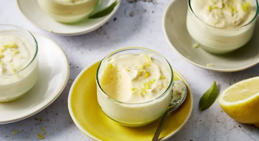 Plusieurs verrines de mousse au citron, garnies de zestes, sur des assiettes claires et jaunes. Un citron coupé est visible à droite.