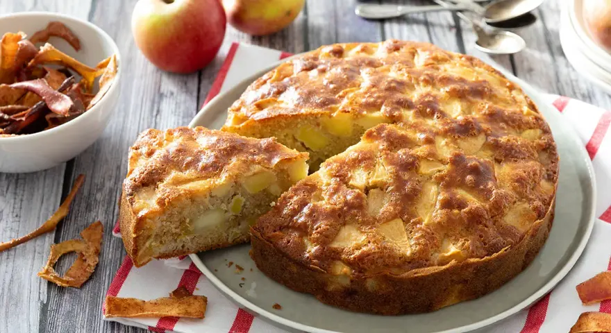Un délicieux gâteau aux pommes, doré et moelleux, avec des morceaux de fruits visibles. Des pommes fraîches et des chips de pommes complètent la scène.