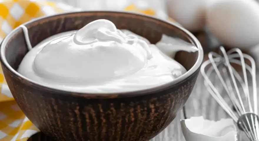 Un bol rempli de crème fouettée, blanc en neige, prêt à être utilisé pour une pâtisserie. Des œufs et un fouet sont visibles à proximité.