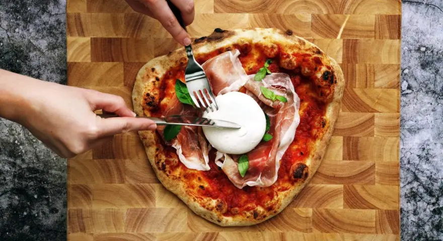 Une pizza burrata avec du prosciutto et du basilic, prête à être dégustée. Des mains coupent la burrata crémeuse.