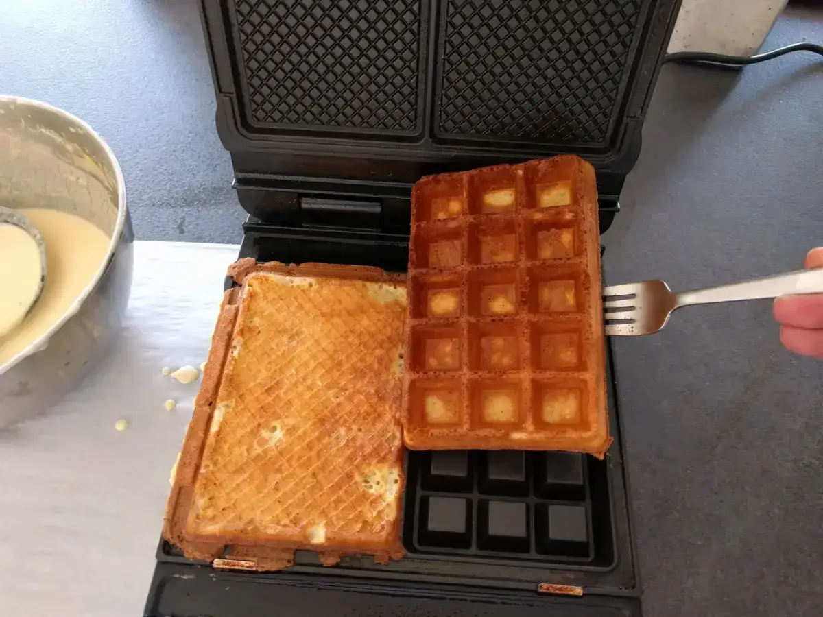 Préparation d'une recette gaufres croustillantes : une gaufre dorée est retirée du gaufrier avec une fourchette, à côté de la pâte.