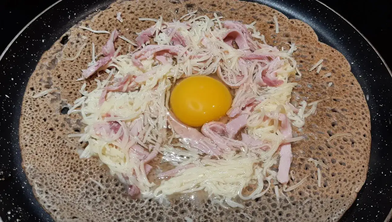 Une galette sarrasin garnie d'un œuf, de jambon et de fromage râpé, prête à être cuite dans une poêle.