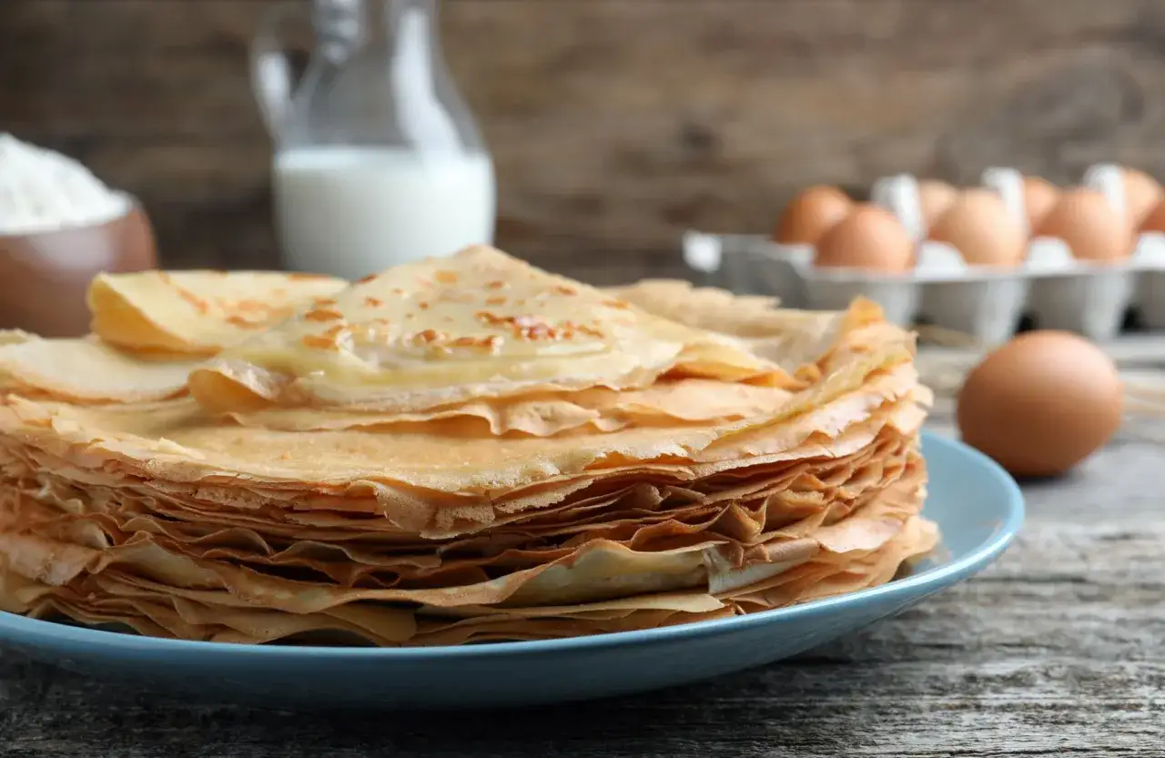 Pile de crêpes dorées sur une assiette bleue, avec des ingrédients pour une recette de crêpe en arrière-plan : farine, lait, œufs.
