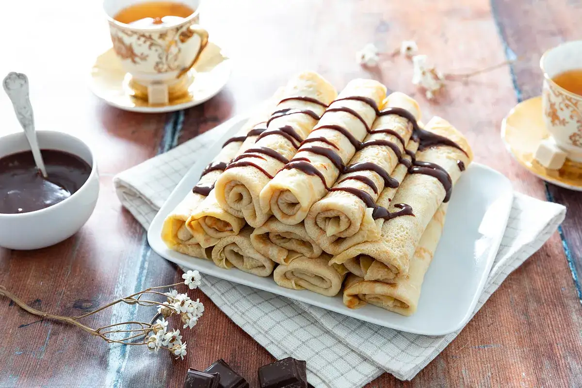 Pile de cr&ecirc;pes moelleuses arros&eacute;es de chocolat, accompagn&eacute;es d'une tasse de th&eacute; et d'une coupelle de sauce chocolat. Une invitation &agrave; la gourmandise.