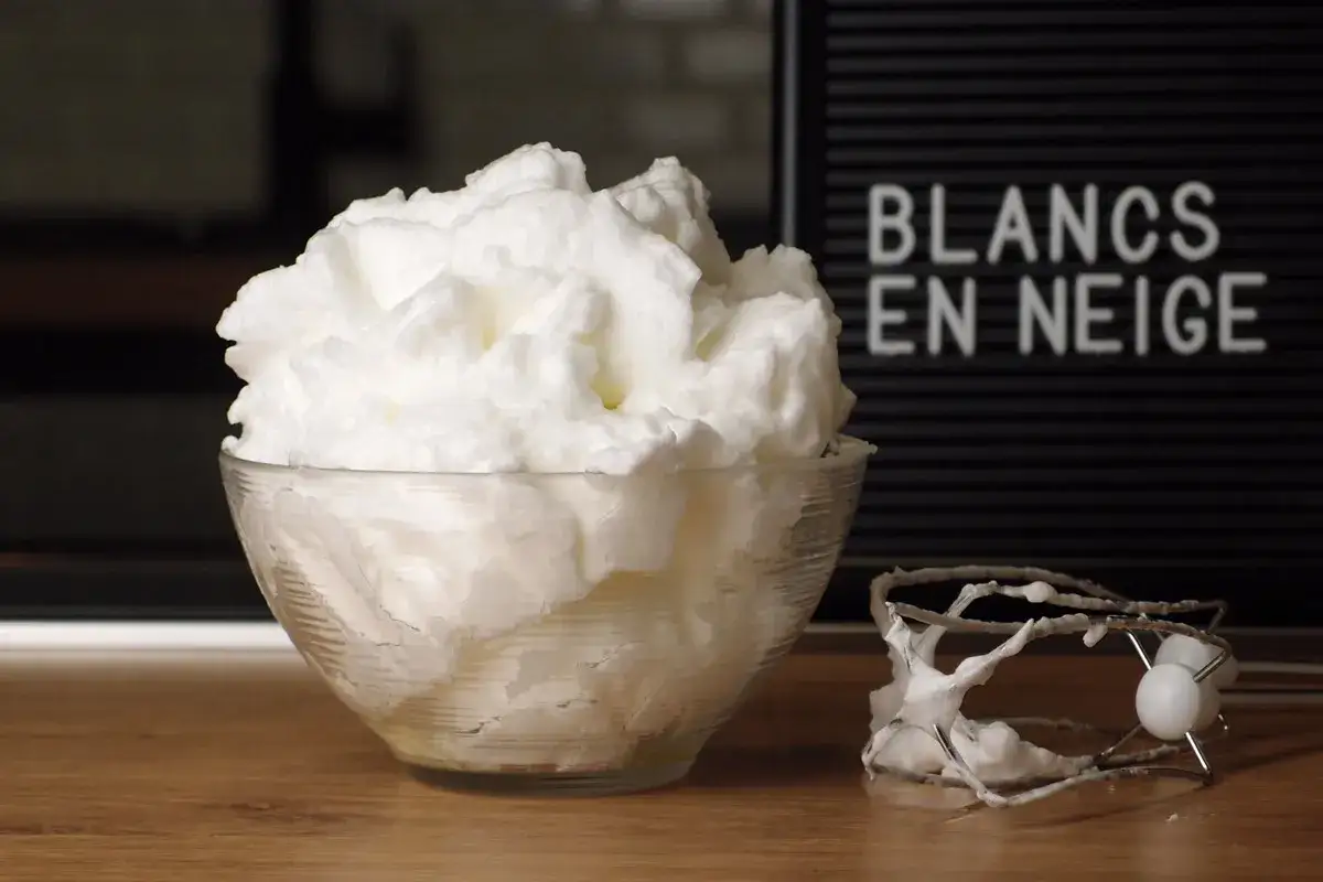 Un bol rempli de blancs en neige aériens, à côté d'un batteur couvert de cette mousse légère.