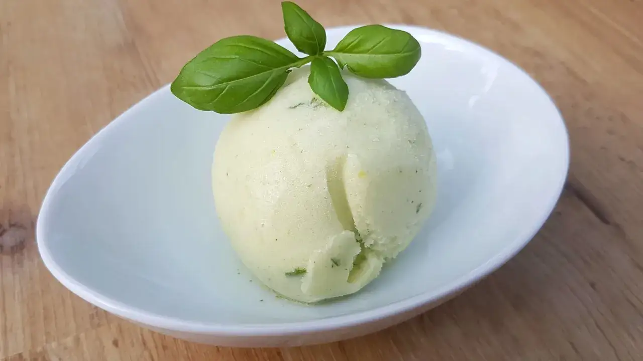Une boule de sorbet citron, garnie de feuilles de basilic frais, repose dans une coupelle blanche sur une table en bois.