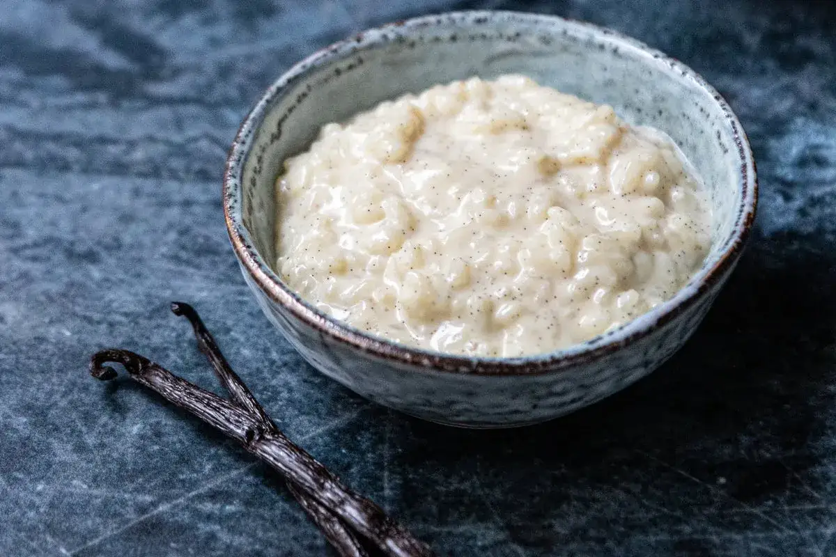 Un bol de riz au lait cr&eacute;meux, parsem&eacute; de points de vanille, avec deux gousses de vanille &agrave; c&ocirc;t&eacute;.