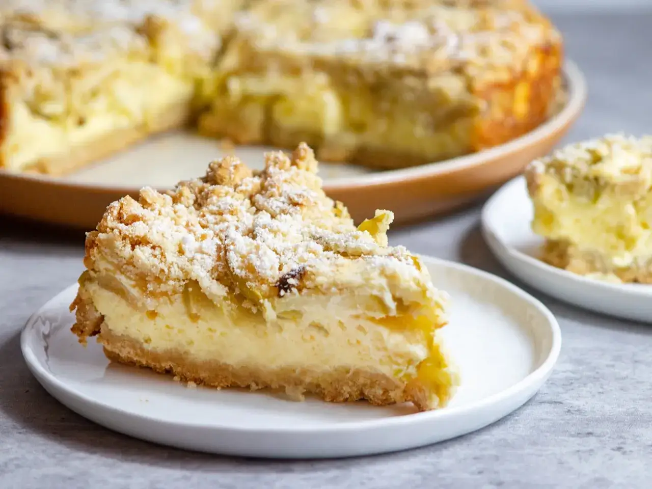Une part de g&acirc;teau mascarpone pommes, saupoudr&eacute;e de sucre glace, sur une assiette blanche. Le reste du g&acirc;teau est visible en arri&egrave;re-plan.