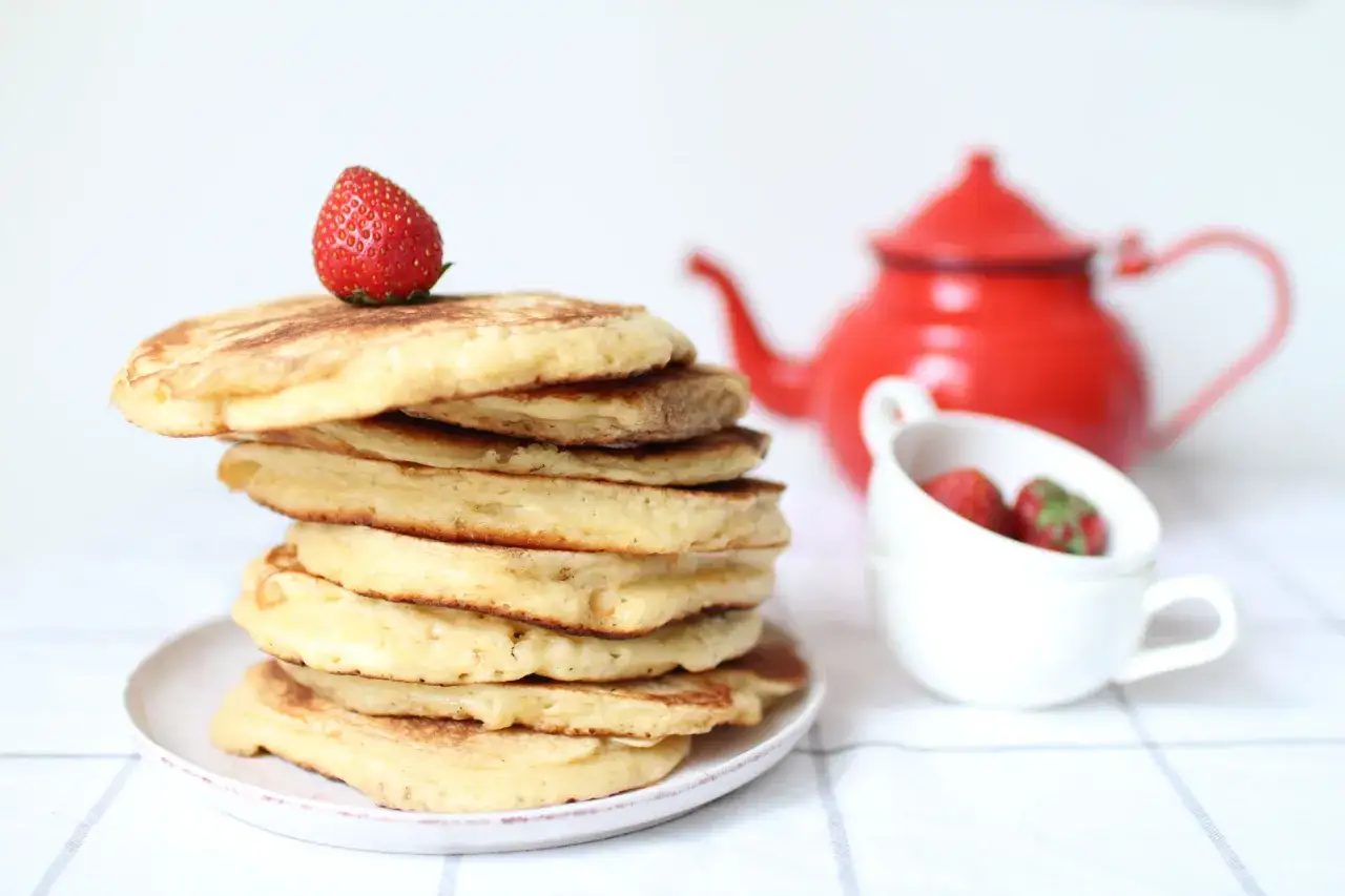 Pile de pancakes sans gluten sur une assiette, garnie d'une fraise. Une th&eacute;i&egrave;re rouge et des fraises dans une tasse sont en arri&egrave;re-plan.