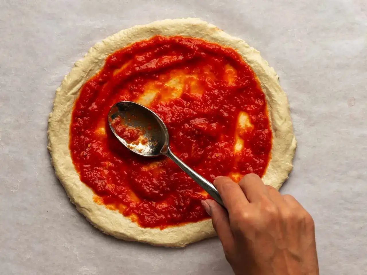 Une main &eacute;tale un coulis de tomates sur une p&acirc;te &agrave; pizza avec une cuill&egrave;re.