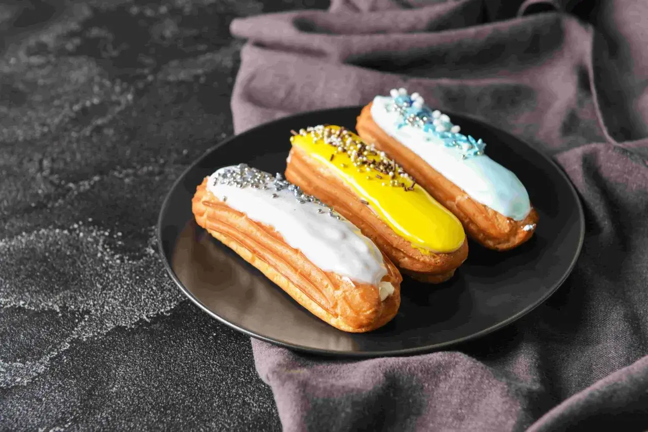 Trois éclairs colorés, faits de pâte à choux, sur une assiette noire. Un blanc, un jaune vif et un bleu ciel, décorés de vermicelles.