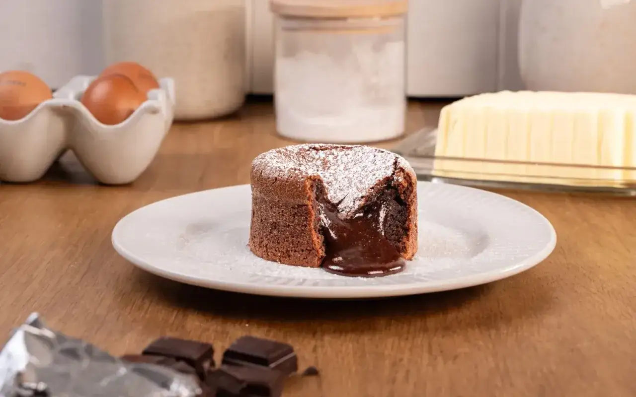 Un d&eacute;licieux coulant au chocolat, saupoudr&eacute; de sucre glace, avec son c&oelig;ur fondant qui coule. Des ingr&eacute;dients de p&acirc;tisserie sont visibles en arri&egrave;re-plan.