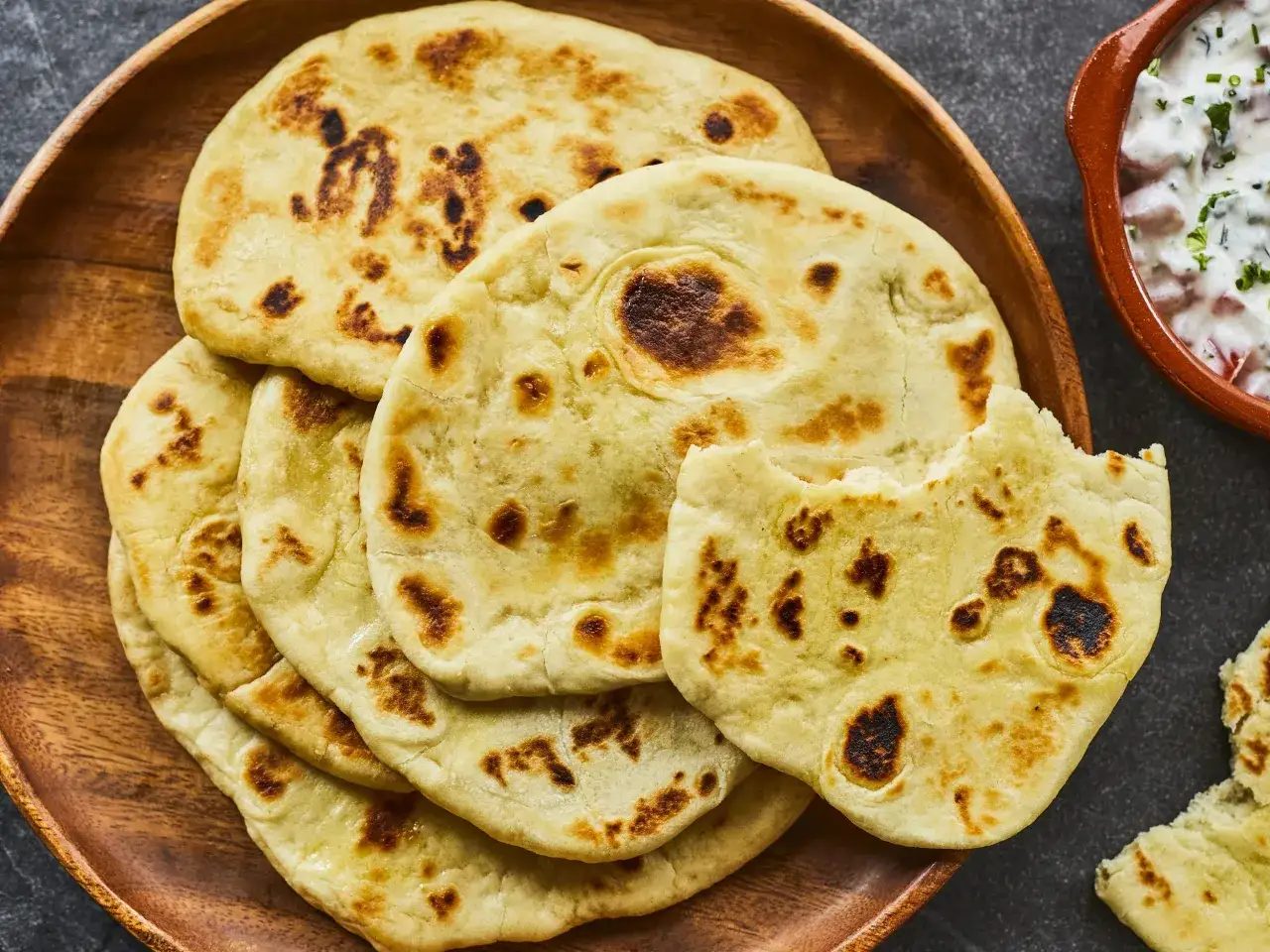 Pile de délicieux cheese naan dorés, certains légèrement brûlés, sur un plat en bois. Un morceau a été mordu.
