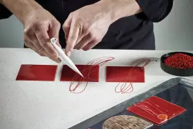 Mains d'un pâtissier créant un décor royal chocolat sur des carrés rouges brillants.