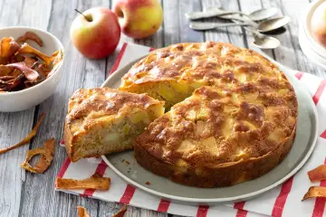 Un délicieux gâteau aux pommes, doré et moelleux, avec des morceaux de fruits visibles. Des pommes fraîches et des chips de pommes complètent la scène.