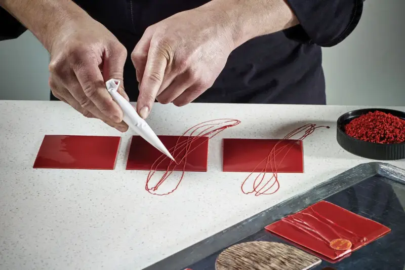 Mains d'un pâtissier créant un décor royal chocolat sur des carrés rouges brillants.